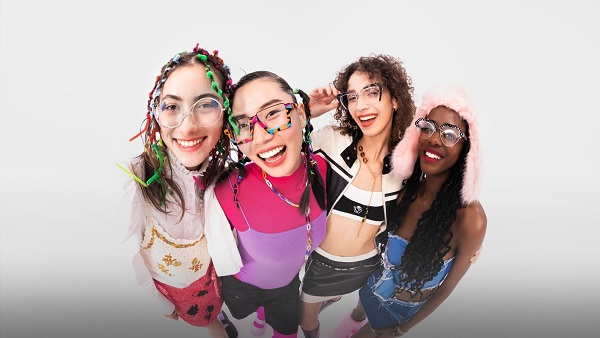 A group of four diverse women in colorful festival outfits, all smiling and wearing unique, prescription-ready rave glasses.