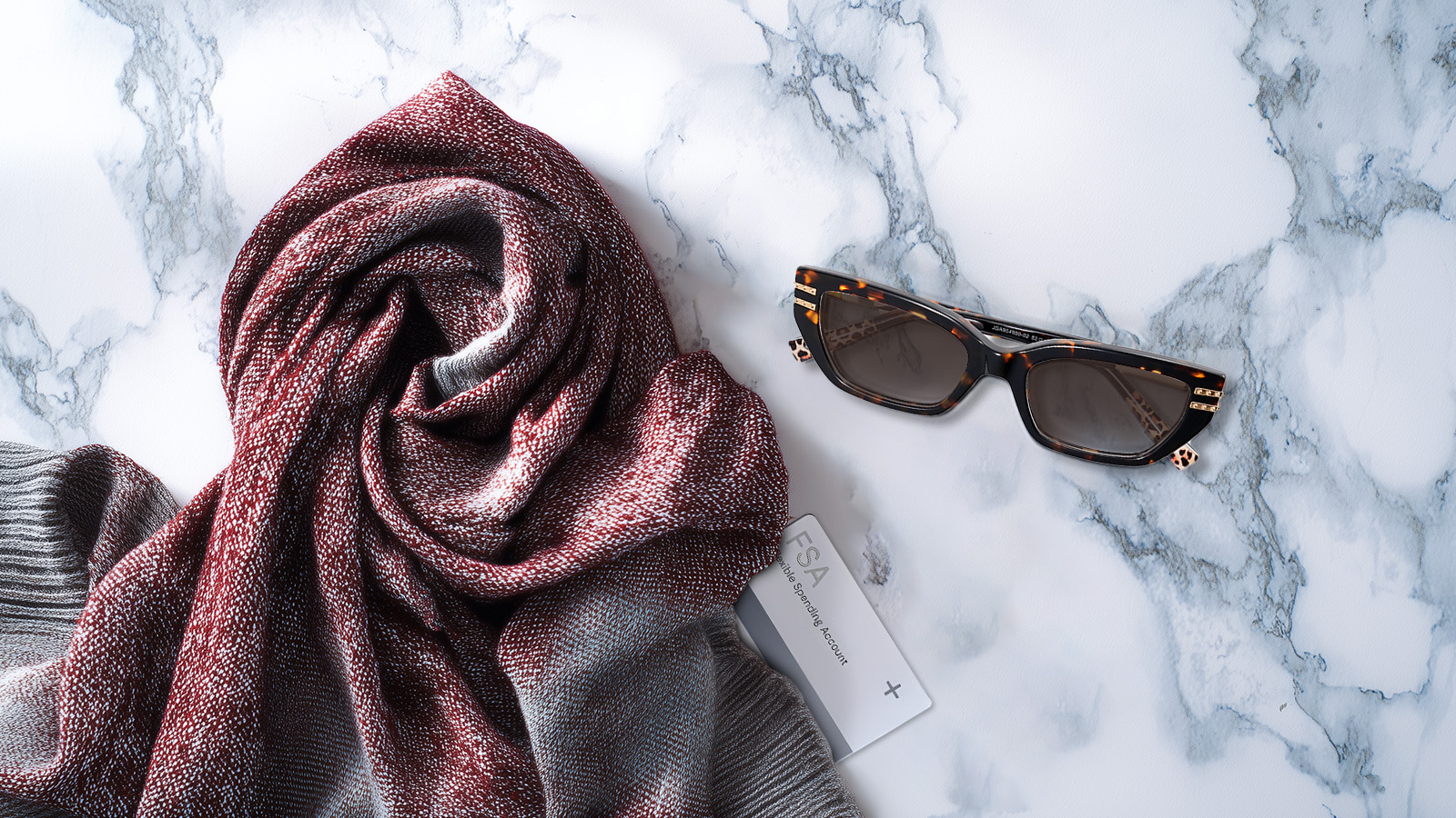 Stylish tortoiseshell sunglasses and an FSA card arranged on a marble surface with a winter scarf, illustrating that sunglasses are a year-round eligible expense.