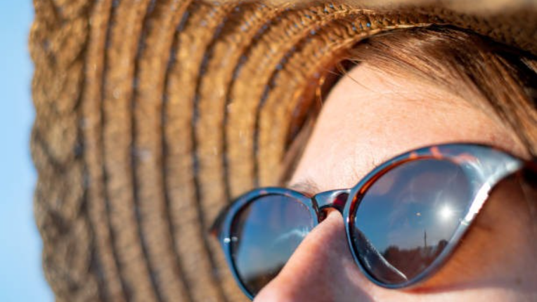 A close-up photograph of a woman outdoors wearing dark sunglasses and a wide-brimmed straw hat to protect against bright sunlight.