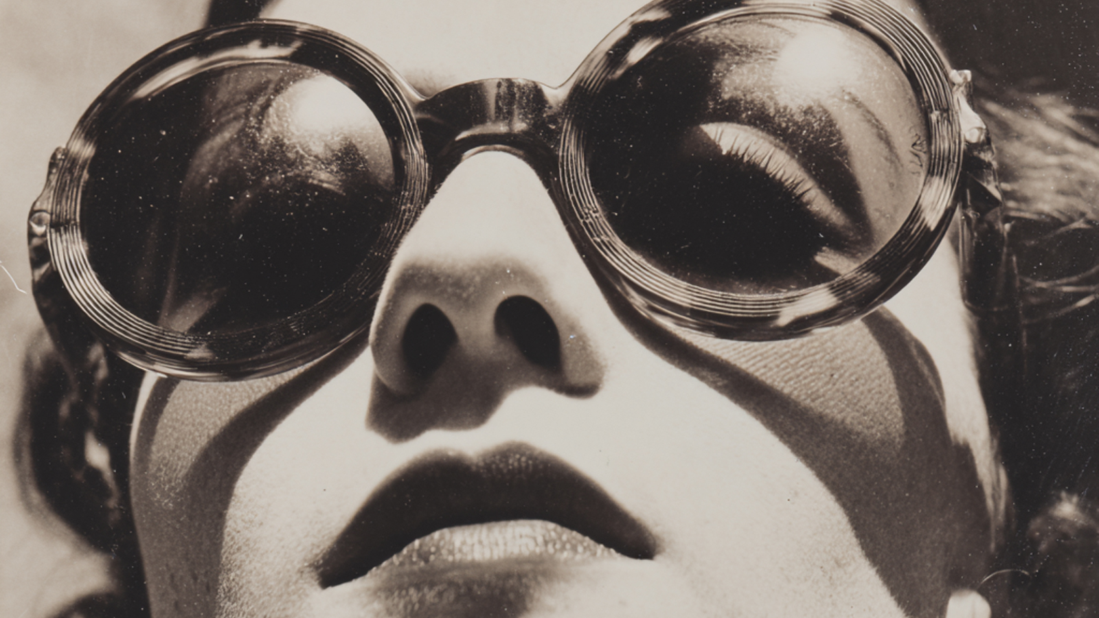 A black and white close-up of a woman wearing large, round, vintage tortoiseshell sunglasses, representing eyewear as a fashion statement.