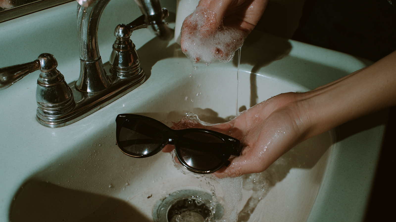 A person rinsing sunglasses under a gentle stream of water, the crucial first step to wash away debris before cleaning.