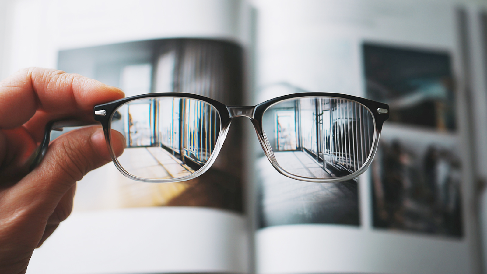 A person holds a pair of glasses over a book, showing how the lenses bring the blurry background into sharp, clear focus.