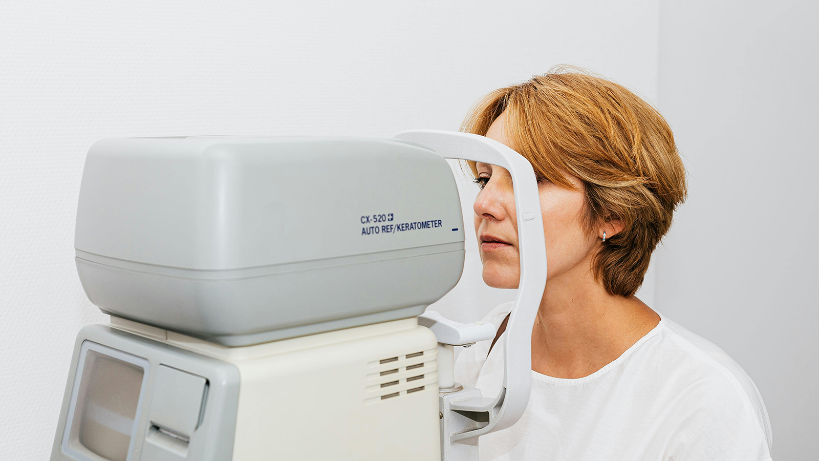 A woman with short, light brown hair looking into an auto ref/keratometer machine during an eye examination, with a neutral white background.