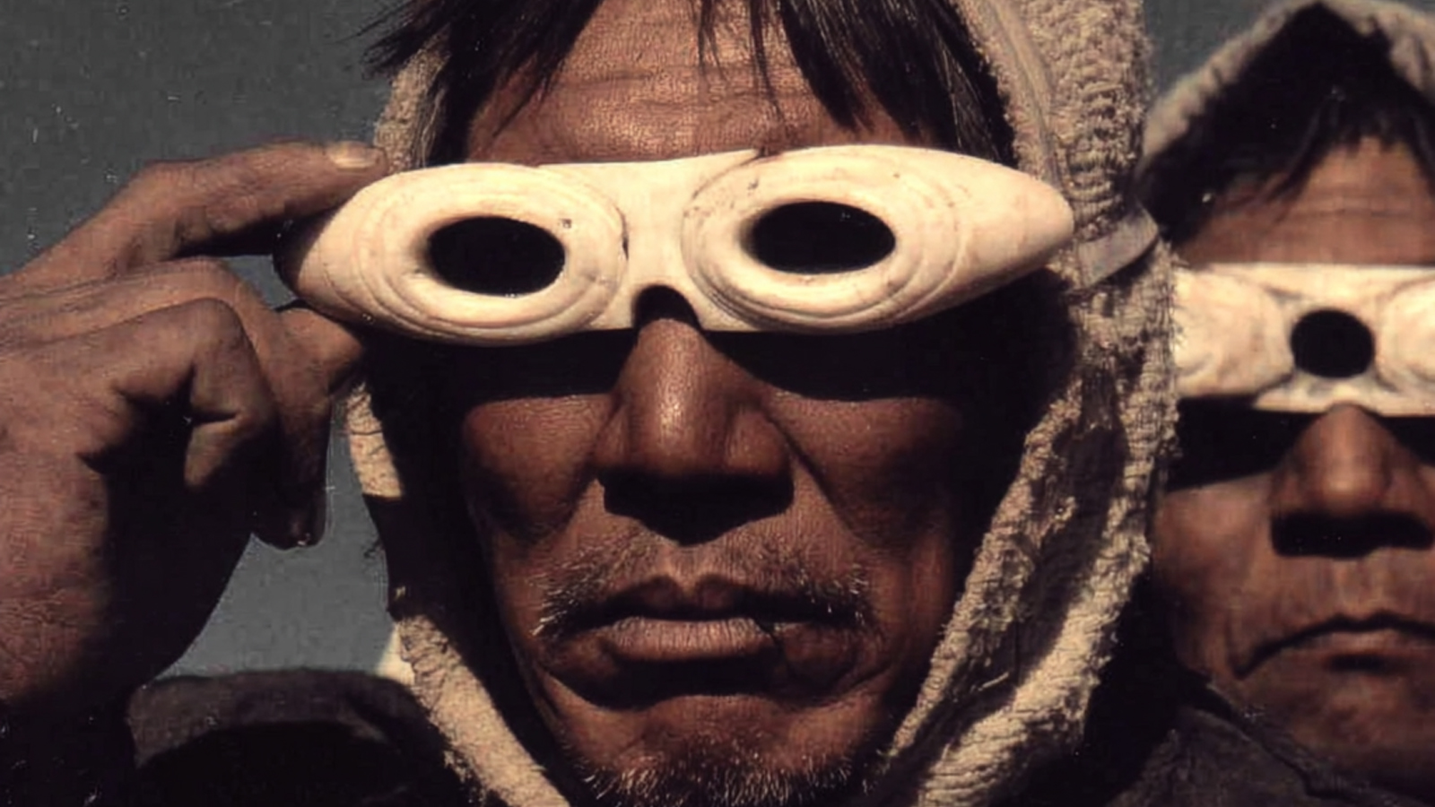 A historical photograph of two Inuit men wearing traditional snow goggles carved from bone to prevent snow blindness.