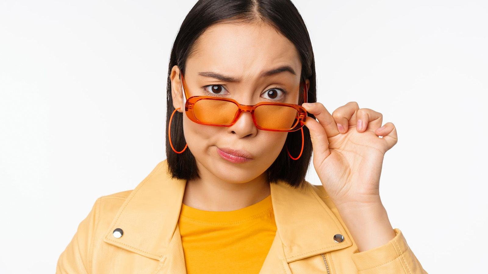 A young woman with a skeptical expression pushes her orange-tinted glasses up her nose with one hand, looking directly at the viewer.