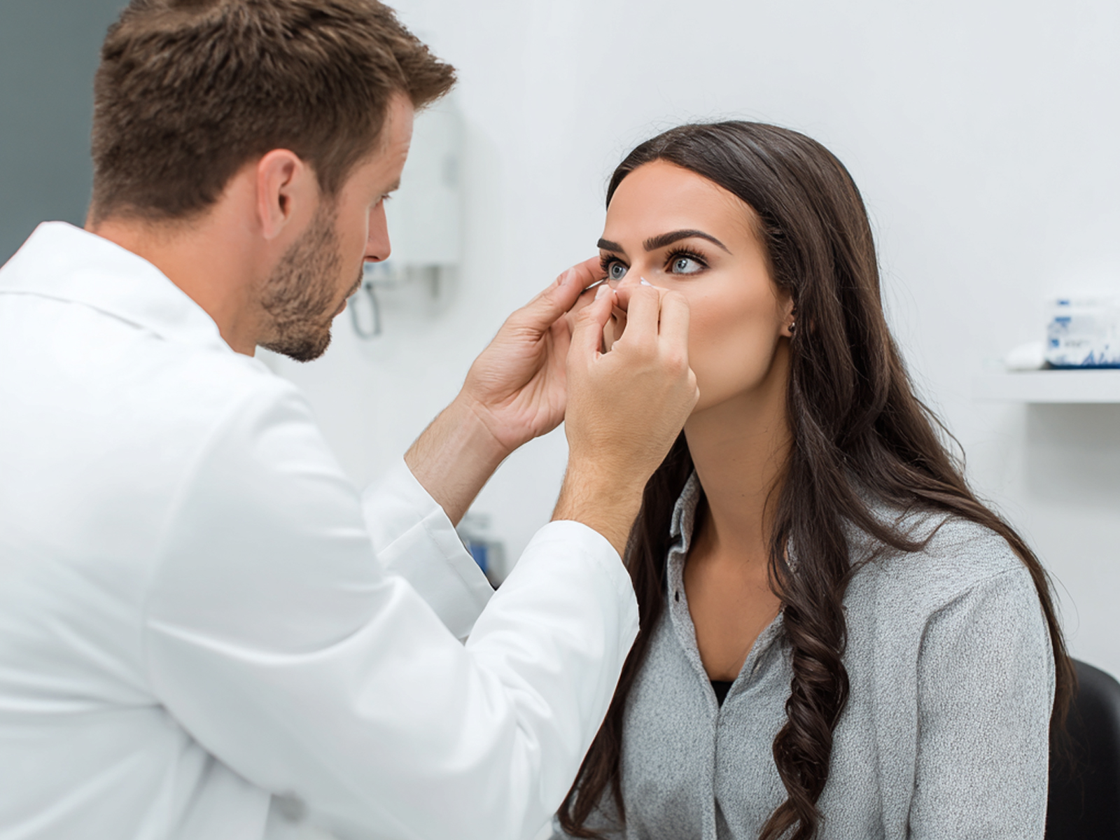 An optometrist in a white lab coat carefully conducts a contact lens fitting for a young woman in a clinical office setting, highlighting the need for professional care when switching from glasses to contacts.