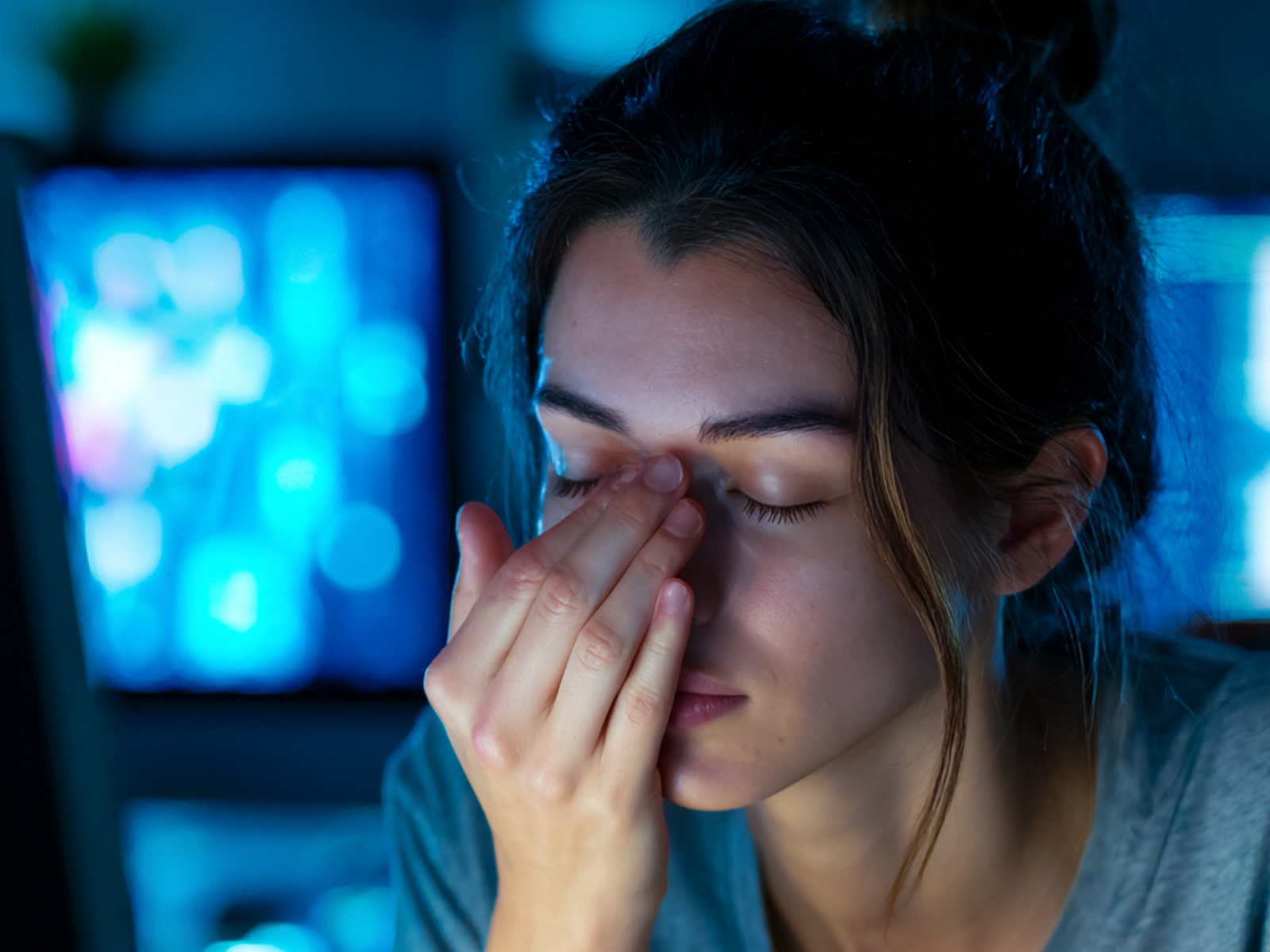A woman experiencing digital eye strain, pinching the bridge of her nose with her eyes closed in front of glowing computer screens in a dark room.